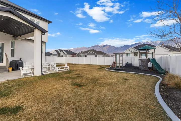 Expansive backyard featuring a covered patio, a manicured lawn, a white privacy fence, and a dedicated play area with a slide and climbing structure