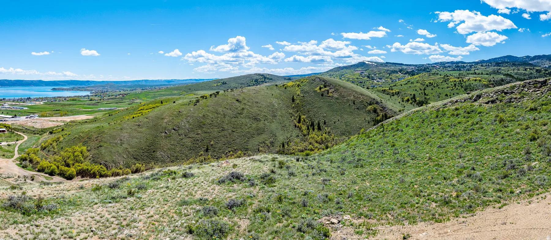 Expansive natural landscape featuring rolling hills, verdant greenery, a distant body of water, and a bright blue sky with scattered clouds
