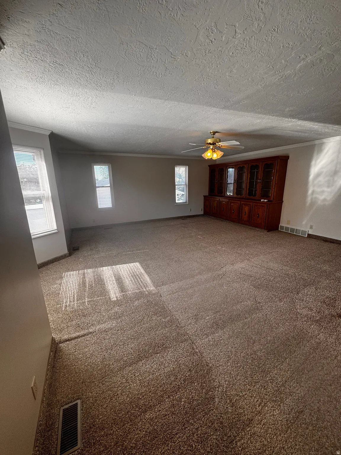 Spacious room featuring neutral carpeting, a built-in wood-finish display cabinet, multiple windows, a ceiling fan with integrated lighting, and crown molding