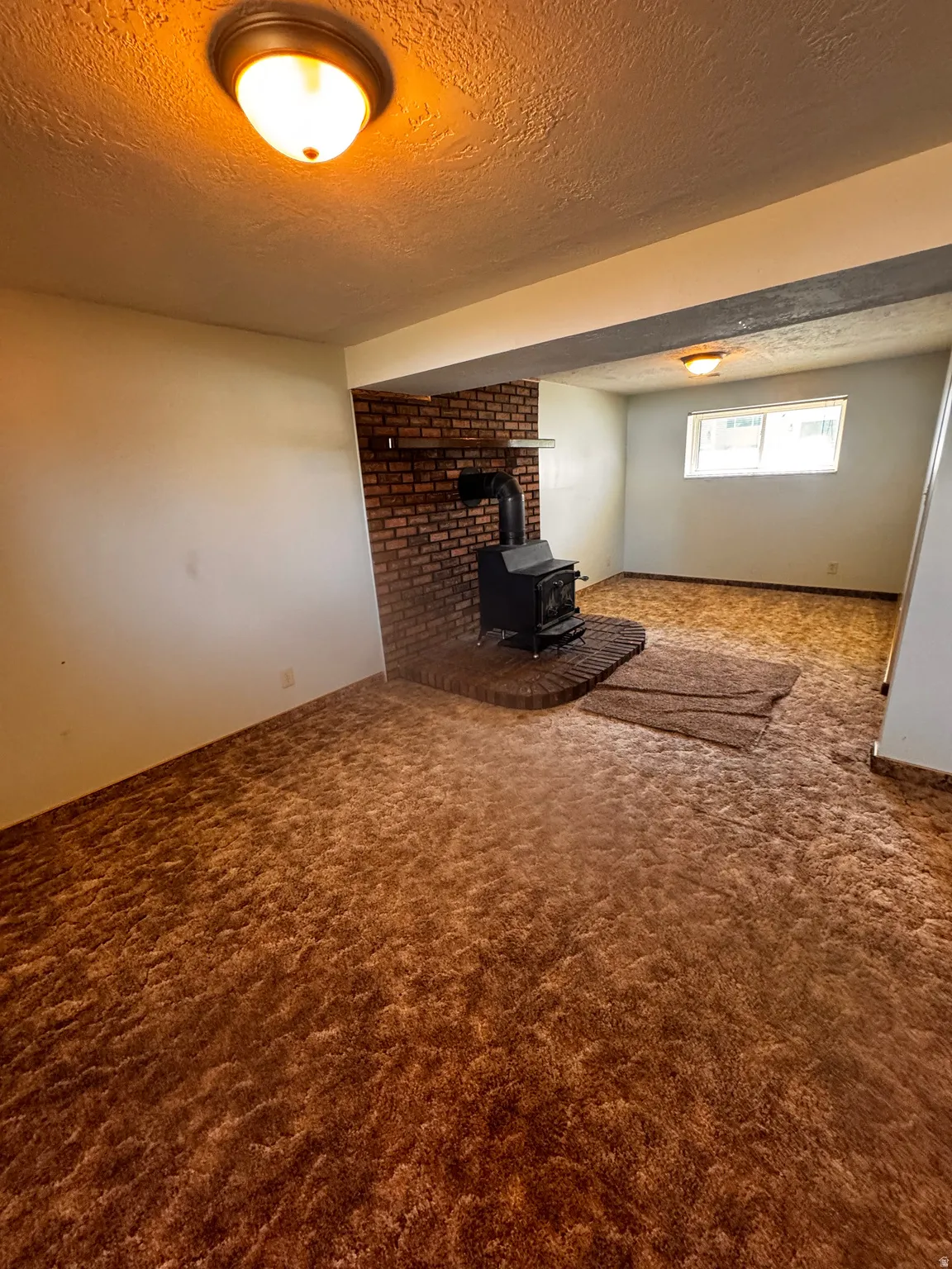 Interior space featuring a brick fireplace with a black wood-burning stove, textured ceiling, flush-mount lighting, and a window with white trim
