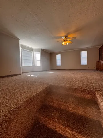 Carpeted living area featuring a bay window with blinds, additional windows with blinds, a ceiling fan with integrated lighting, and crown molding