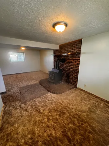 Spacious room featuring a brick accent wall with a wood-burning stove, brown patterned carpet, and a flush mount ceiling light