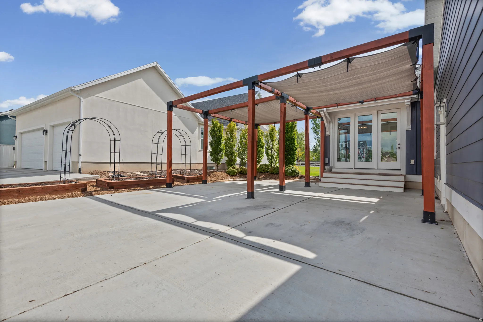Expansive concrete patio with a large wooden pergola featuring a removable shade cover, dark blue horizontal siding, and a white entry door. Additional parking for 3 cars in front of pergola