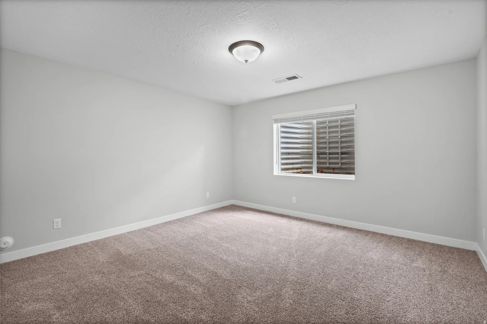Basement ADU bedroom. Neutral wall colors, carpet flooring, a single window with blinds, and an overhead light fixture