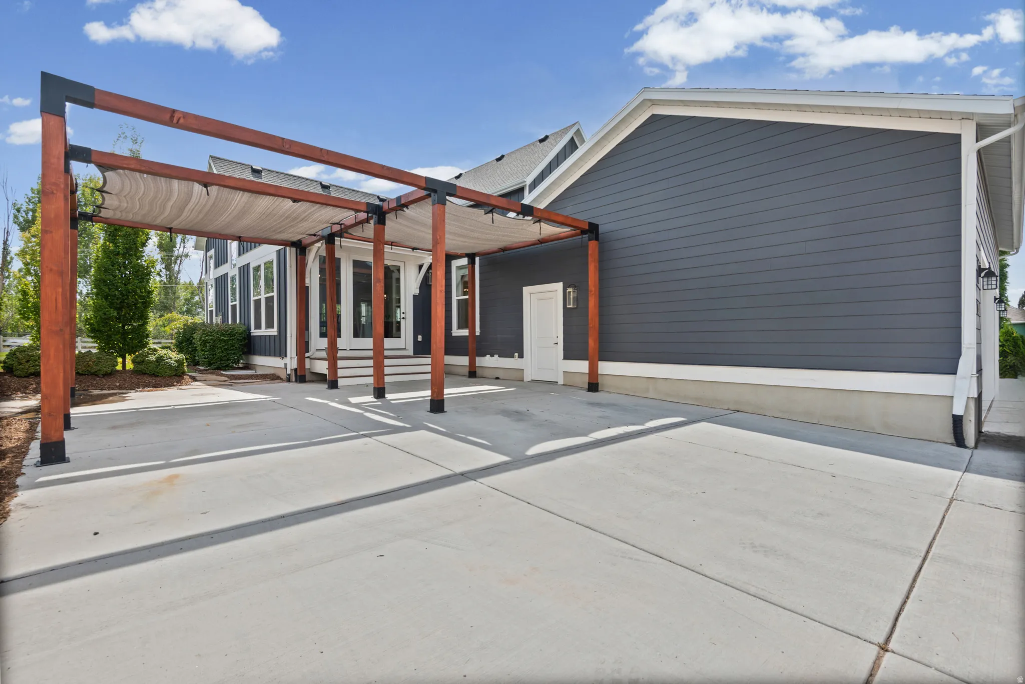 Expansive concrete patio with a large wooden pergola featuring a removable shade cover, dark blue horizontal siding, and a white entry door. Additional parking for 3 cars in front of pergola