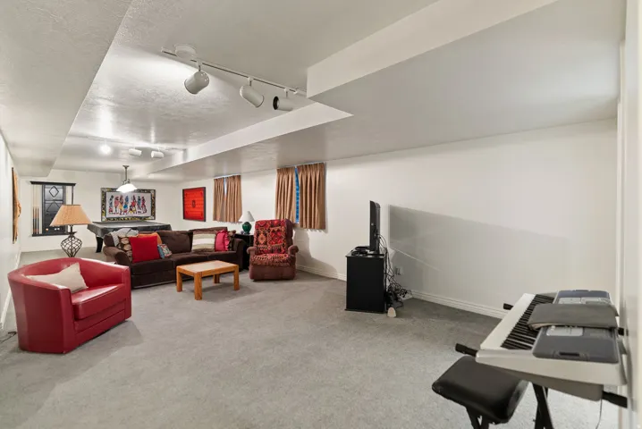 Spacious lower-level room featuring light gray carpeting, track lighting, ceiling-mounted light fixtures, white walls, and white baseboards