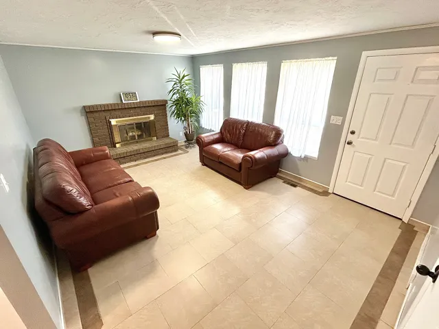 Living area featuring a brick fireplace with a brass firebox, light-toned tile flooring, three large windows, and a six-panel entry door