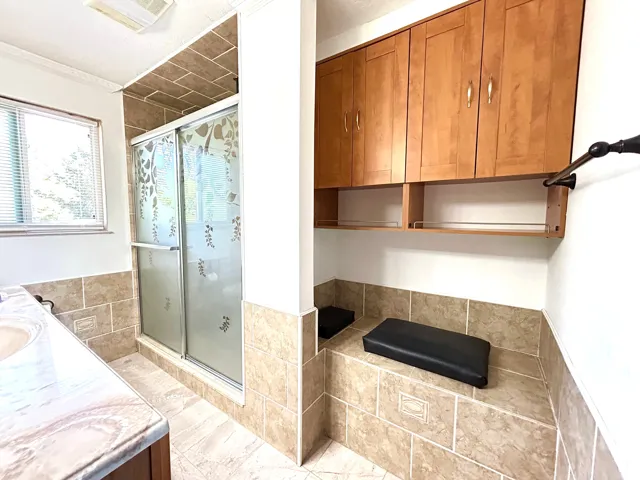 Tiled shower enclosure with sliding frosted glass doors, integrated tiled bench seating, wood-finish cabinetry, and a window with blinds