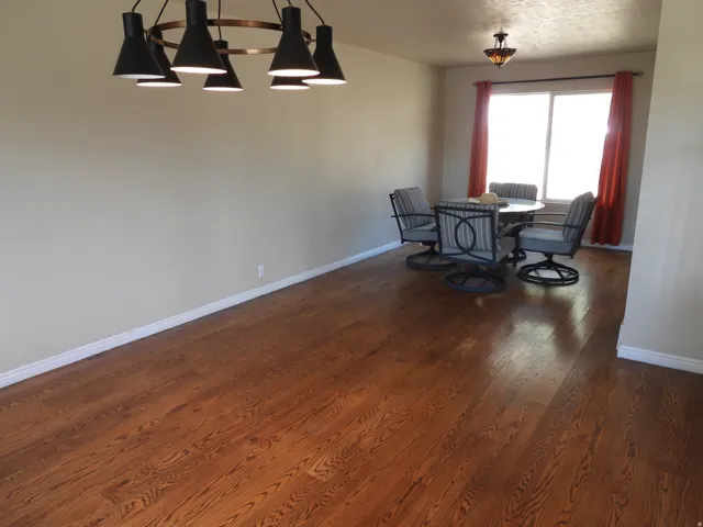 Expansive room featuring rich wood-finish flooring, a modern multi-bulb chandelier, a large window providing ambient light, and white baseboards
