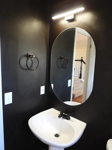 Pedestal sink with black hardware, complemented by a contemporary oval mirror and linear wall-mounted light fixture