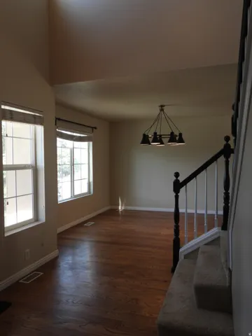 Open-concept living space with dark wood-finish flooring, a contemporary six-light chandelier, dual windows, high ceilings, and a carpeted staircase with a black and white banister