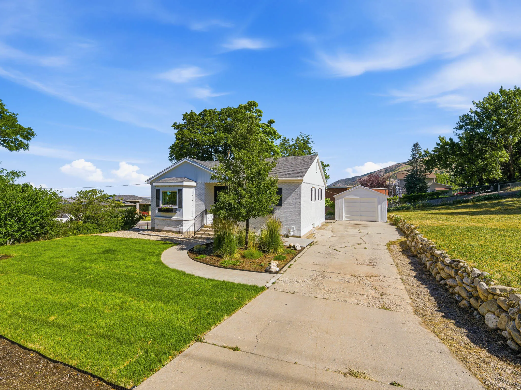 White exterior featuring a bay window with flower box, a detached garage, a stone retaining wall, and mature landscaping