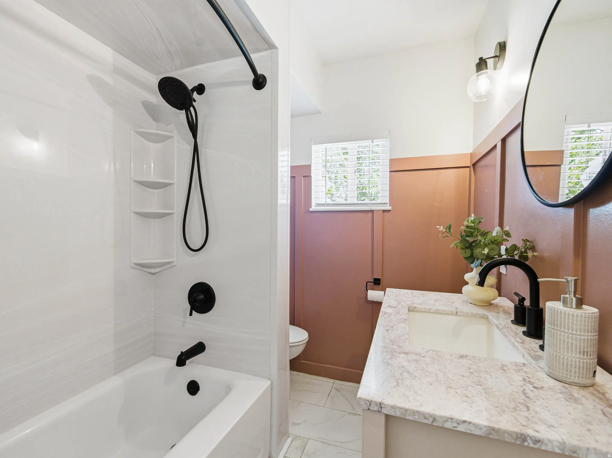 Bathroom featuring a white bathtub with a black shower head and fixtures, a light-colored vanity with an integrated sink, a round mirror, and a window with blinds