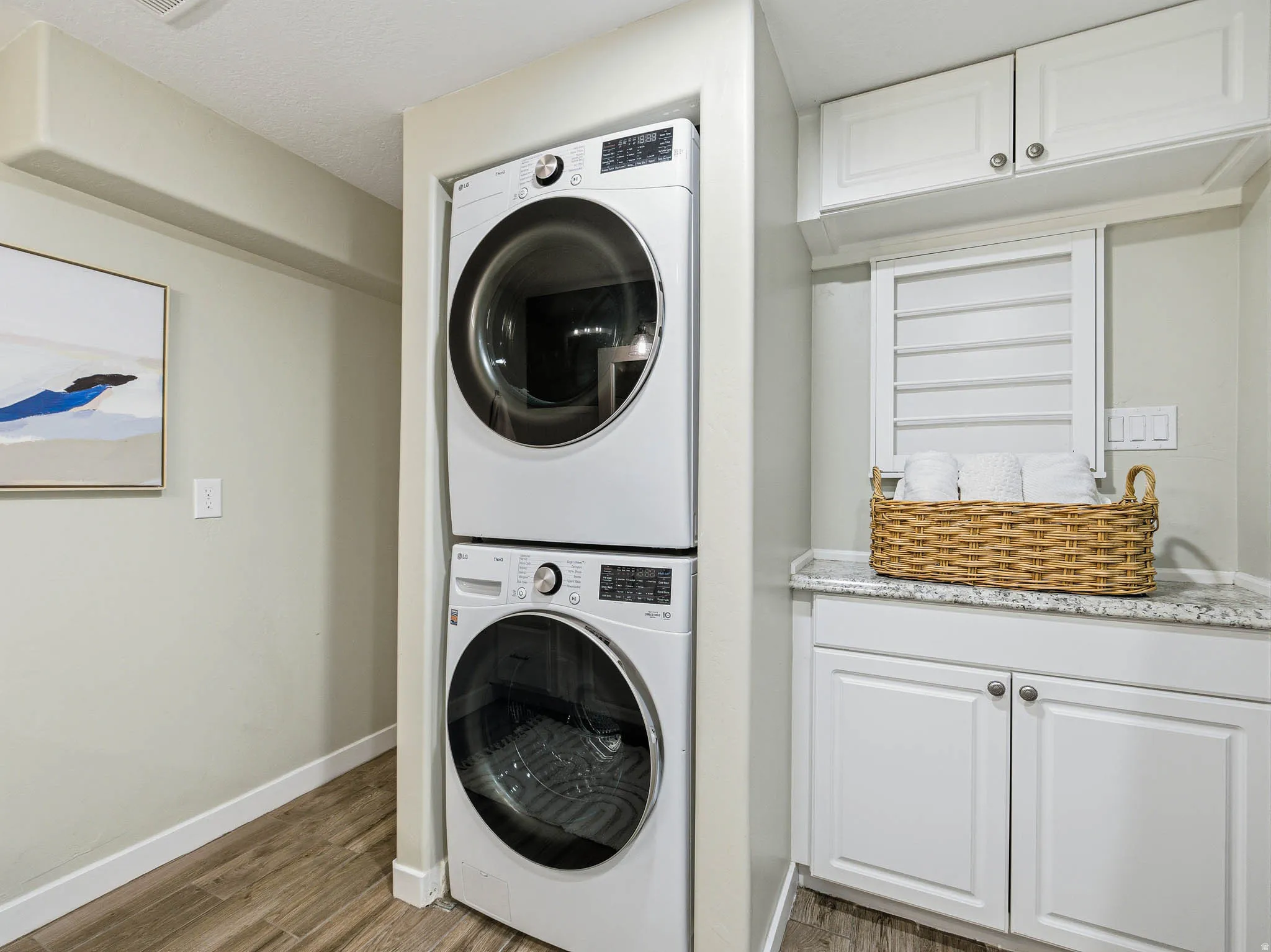 Dedicated laundry area featuring stacked front-load machines, wood-finish flooring, white cabinetry, and a light-toned countertop