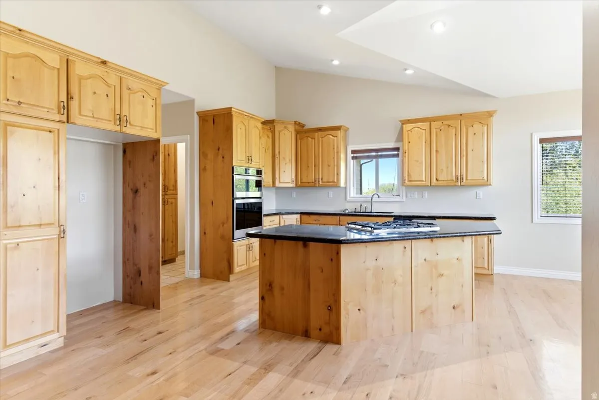 Open-concept kitchen featuring light wood-finish cabinetry, a central island with a dark countertop and built-in stovetop, stainless steel double wall ovens, and light wood-finish flooring