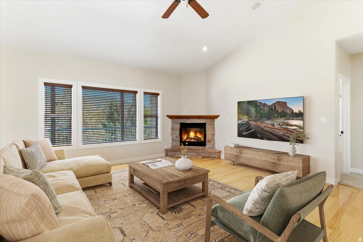 Vaulted ceilings highlight this living space, featuring a stone-clad fireplace with a wood mantle, blonde wood-finish flooring, and a triple window assembly with horizontal blinds.Virtually staged.