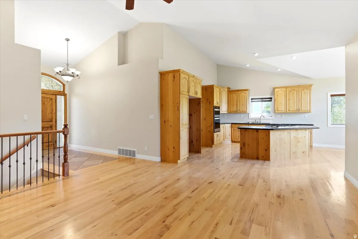 Open-concept living space featuring high vaulted ceilings, light wood-finish flooring, and a kitchen with warm-toned cabinetry, dark countertops, and a central island with a built-in cooktop