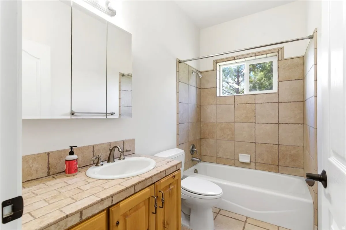 Bathroom featuring an integrated bathtub and shower with tiled surround, a window, and a vanity with a tile countertop, an oval sink, and a mirrored medicine cabinet