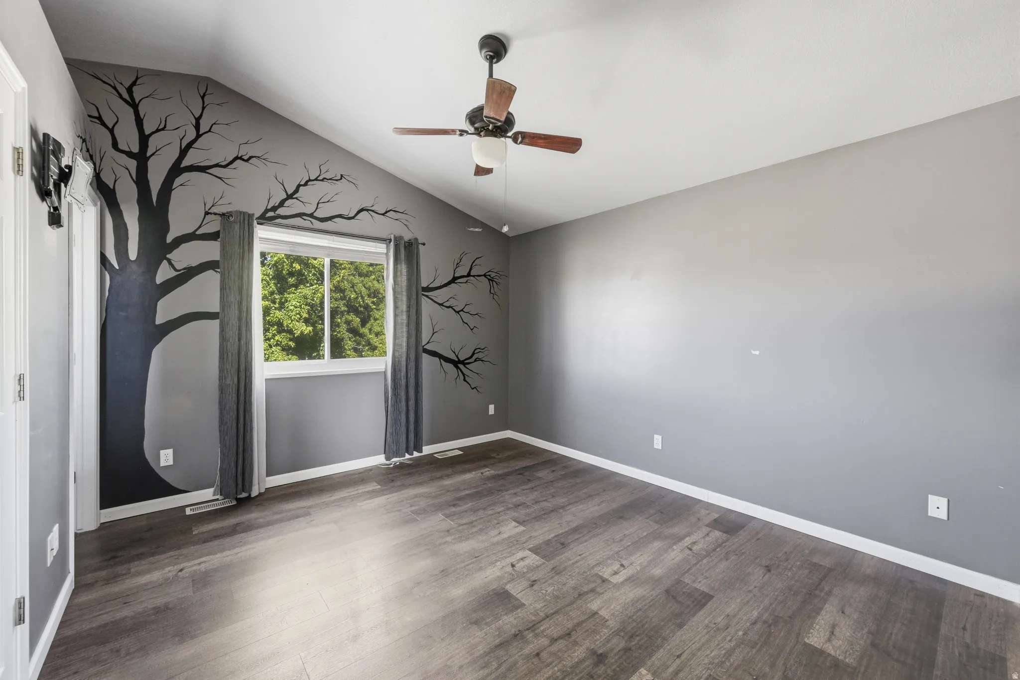 Vaulted ceiling with a ceiling fan, wood-finish flooring, and a window with exterior tree views