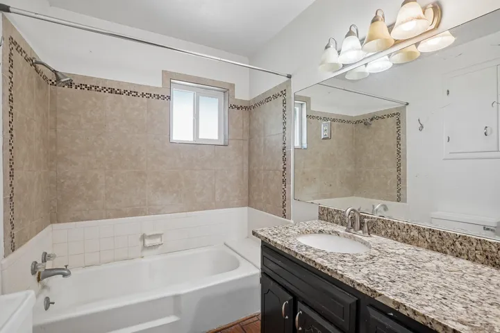 Bathroom featuring a built-in tub with tile surround, a window over the tub, a vanity with a granite-style countertop, an undermount sink, and an expansive mirror