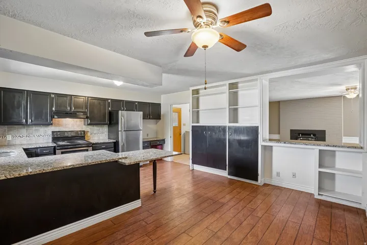 Open-concept living area with wood-finish flooring, a ceiling fan with light fixture, and a kitchen featuring dark cabinetry, stainless steel appliances, and light-toned countertops