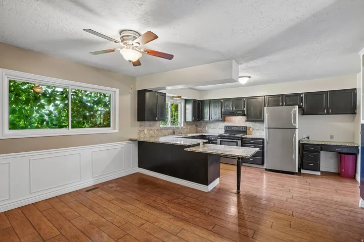 Open-concept kitchen and dining area featuring wood-finish flooring, a ceiling fan, and a large window overlooking lush greenery