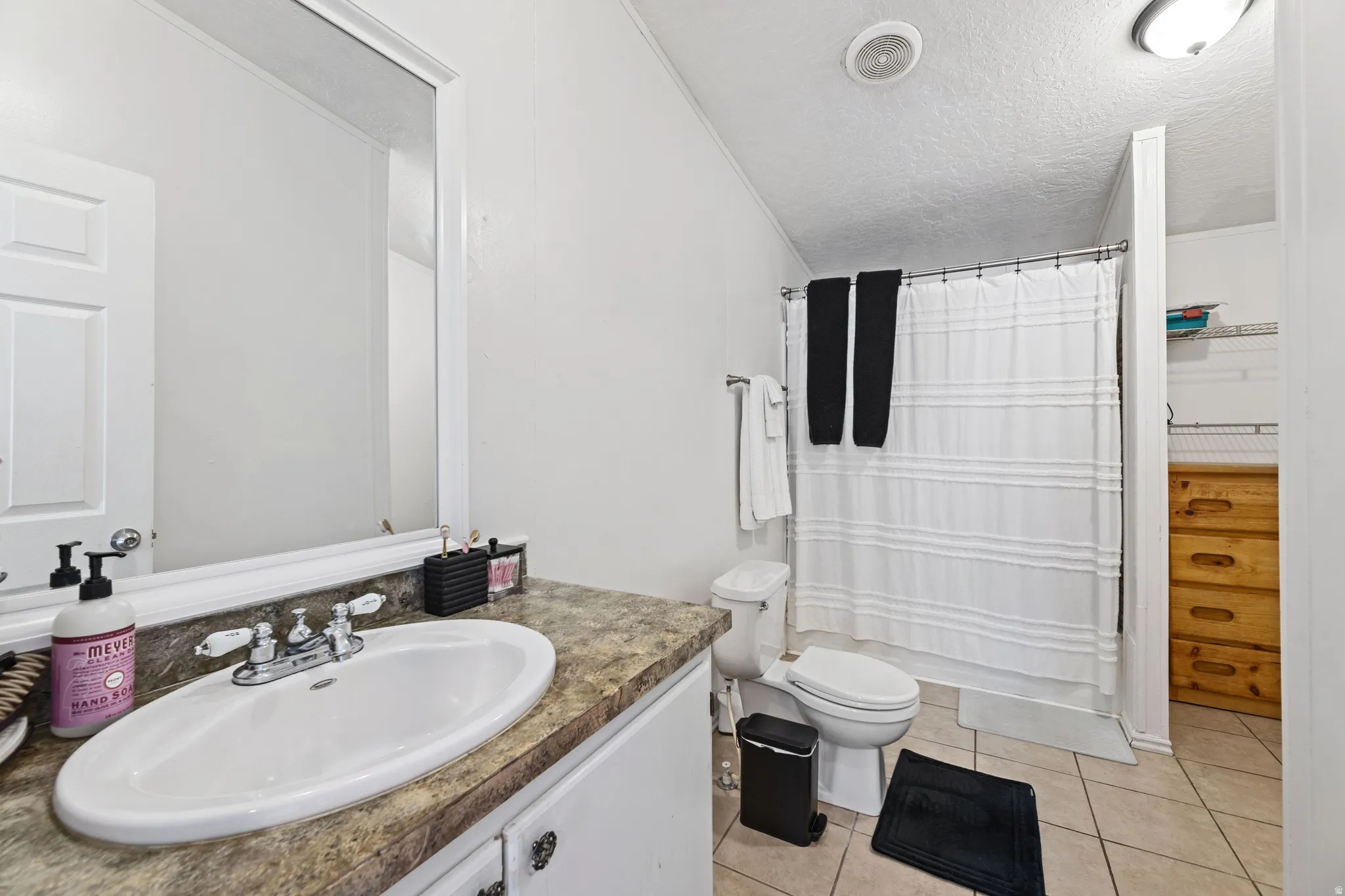 Bathroom featuring a single vanity with a laminate countertop, an oval undermount sink, a shower-tub combination with a white shower curtain, tiled flooring, and an adjacent built-in storage area with shelving and wood-finish drawers