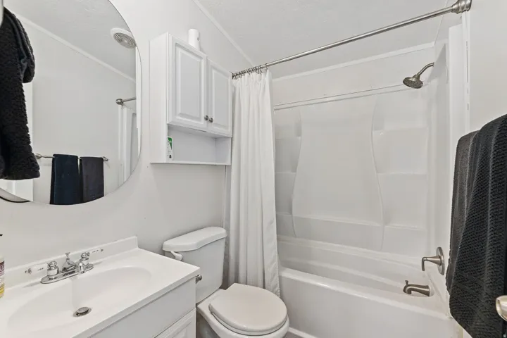 Bathroom featuring a white vanity with an integrated sink, a circular mirror, a white toilet, a shower-over-tub combination with a white surround, and a wall-mounted storage cabinet
