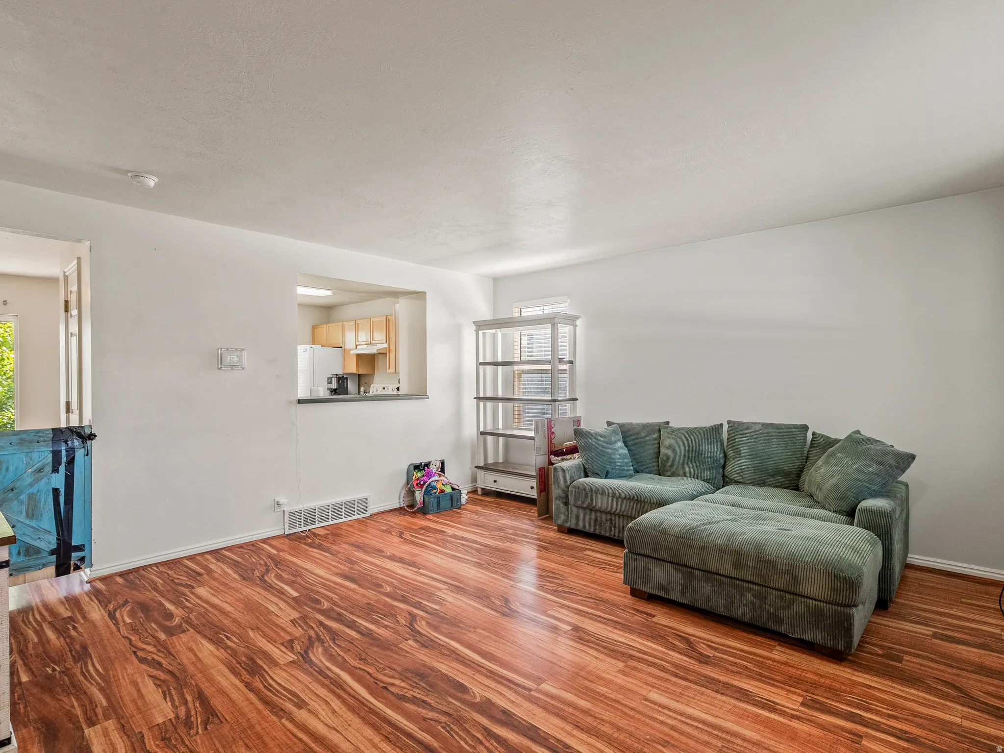 Expansive open-concept living area featuring wood-finish flooring, a pass-through window to the kitchen, white painted walls, and a built-in shelving unit