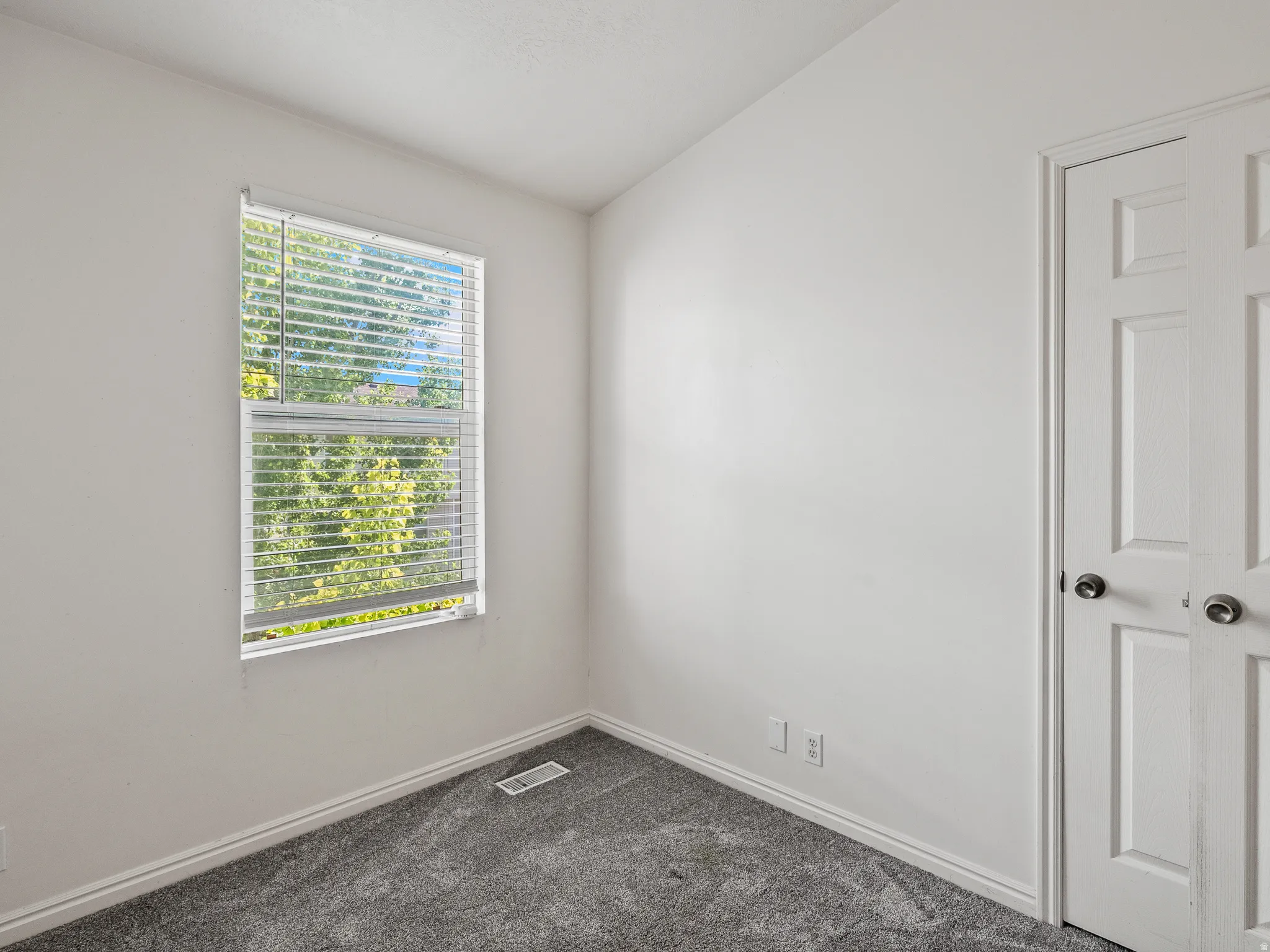 Room featuring a double-pane window with horizontal blinds, gray carpeting, white painted walls, white baseboards, and a white bi-fold closet door with dark hardware