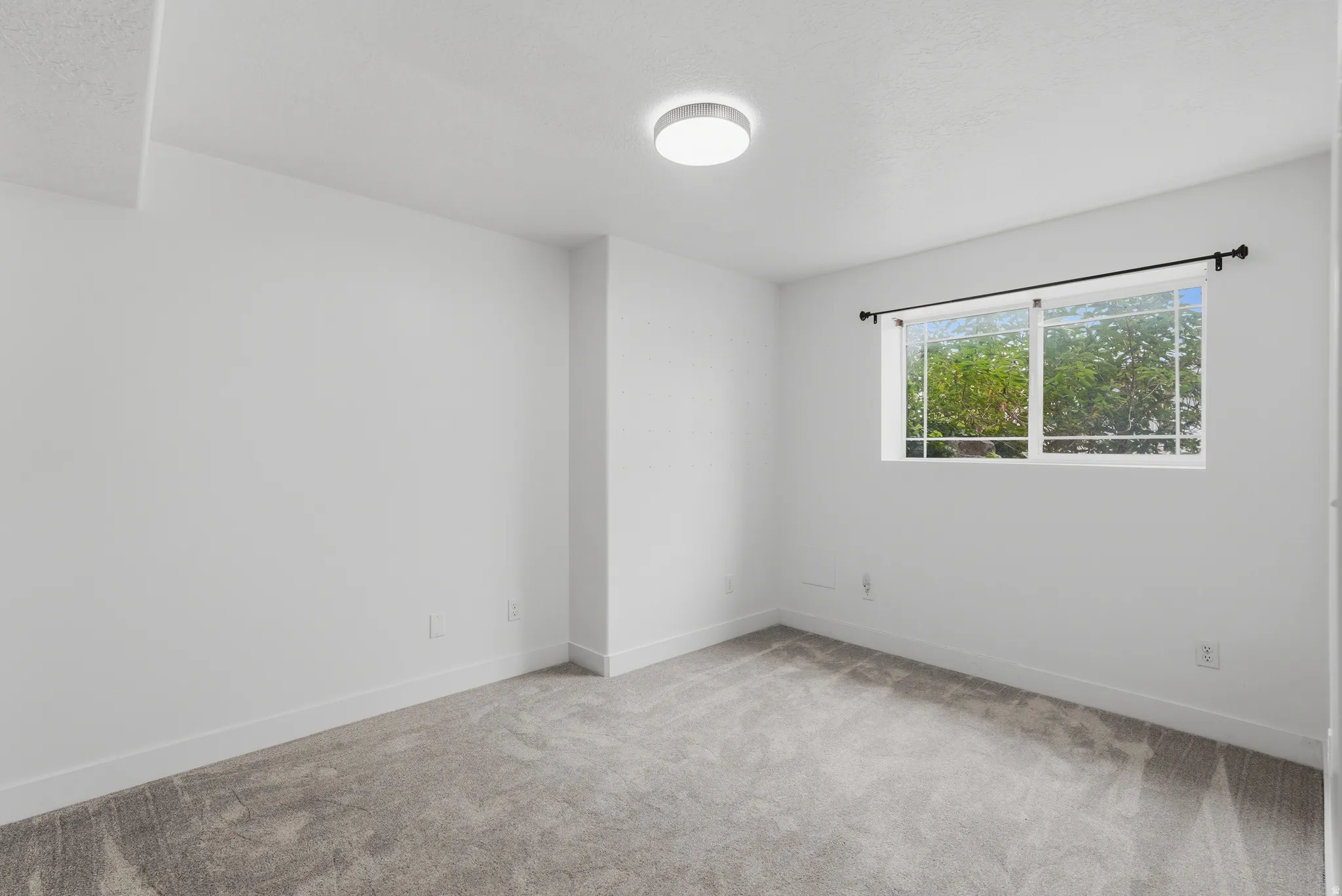 Spacious room featuring light gray carpeting, crisp white walls, a contemporary flush-mount ceiling light, and a large window with black curtain rods