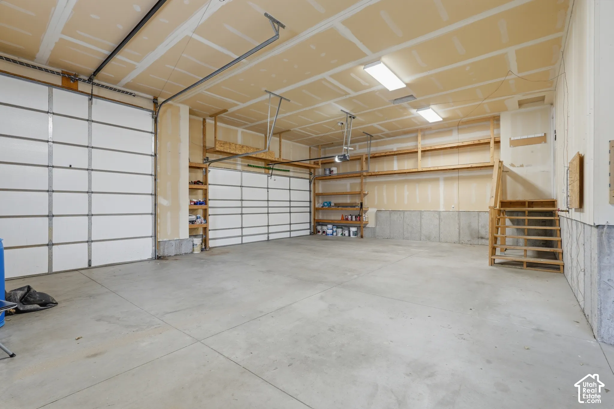 Spacious garage featuring two sectional garage doors, concrete flooring, exposed ceiling joists, built-in wooden shelving, and fluorescent lighting fixtures