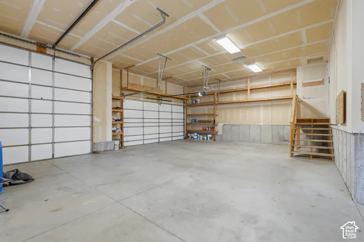 Spacious garage featuring two sectional garage doors, concrete flooring, exposed ceiling joists, built-in wooden shelving, and fluorescent lighting fixtures