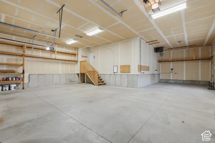 Spacious garage featuring a concrete floor, exposed wood shelving, an interior stairway with wood railings, fluorescent ceiling lighting, and exposed ceiling joists