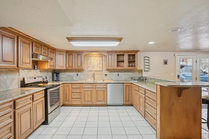 Spacious kitchen featuring light wood cabinetry, stone countertops, a tile backsplash, stainless steel appliances, and white tile flooring