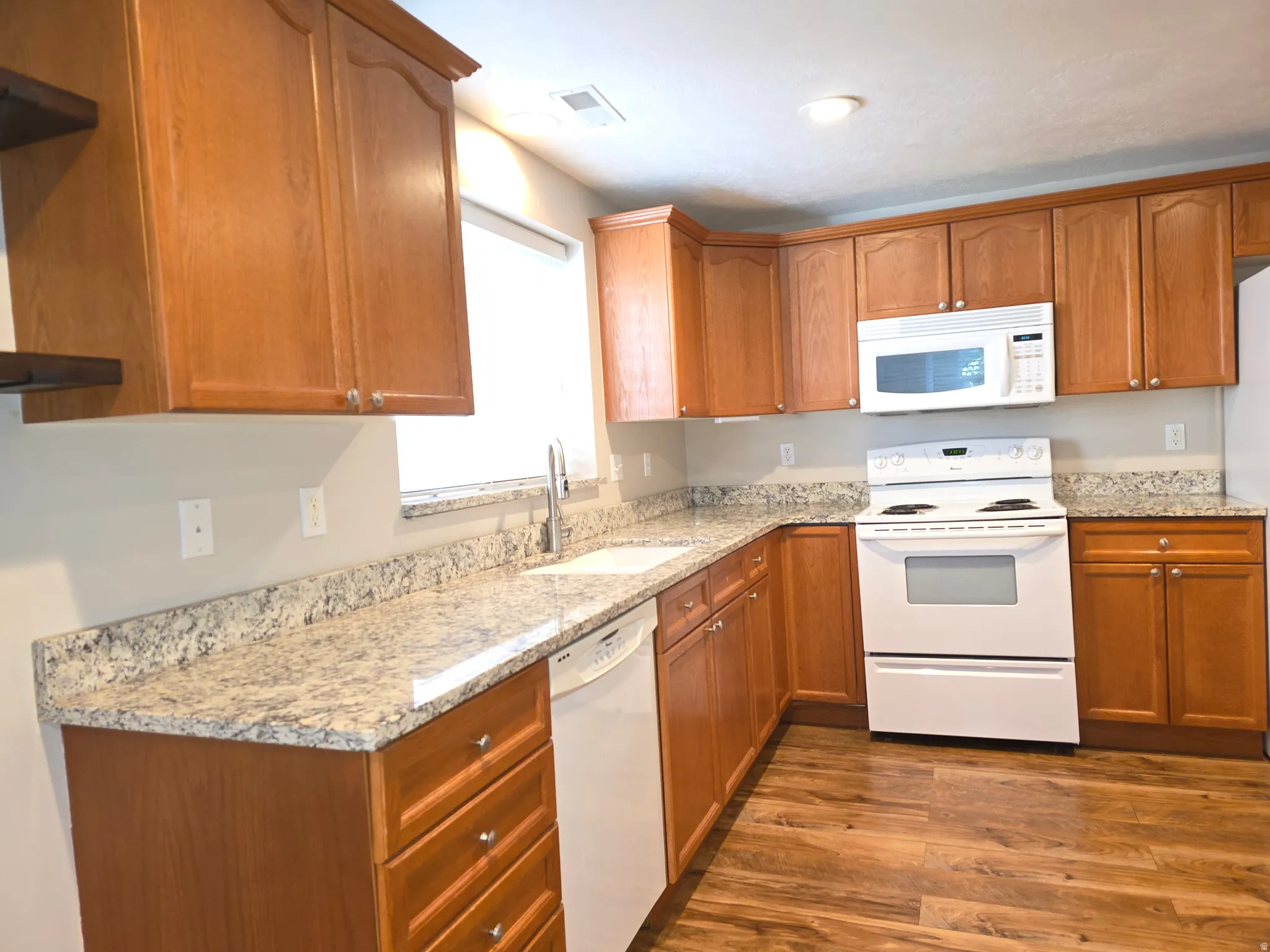 Kitchen featuring wood-finish cabinetry, light-toned countertops, white appliances, a stainless steel sink, and wood-finish flooring