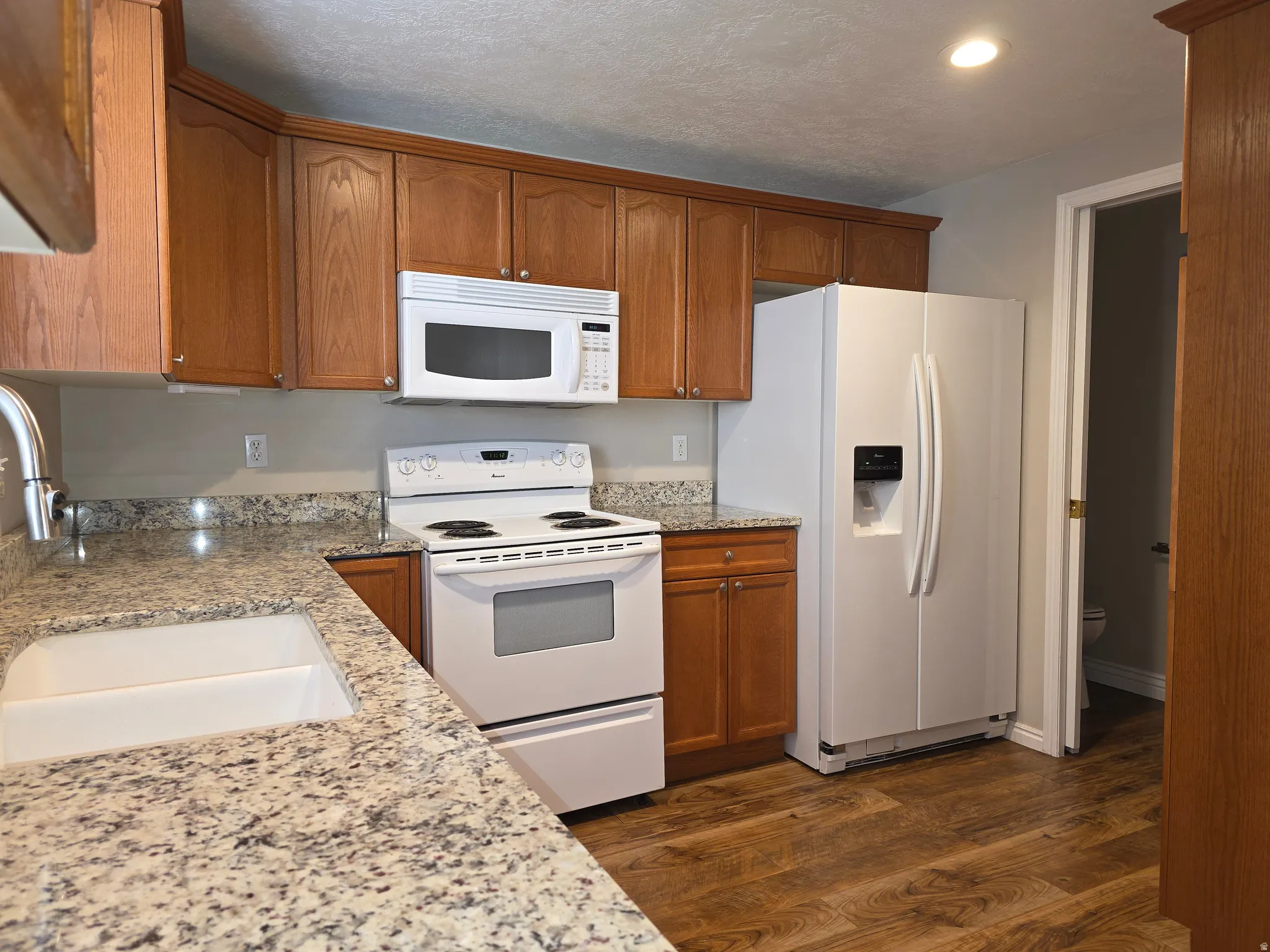 Kitchen featuring a white drop-in sink, speckled countertops, wood-finish flooring, wood cabinetry, and white appliances including a refrigerator, range, and microwave