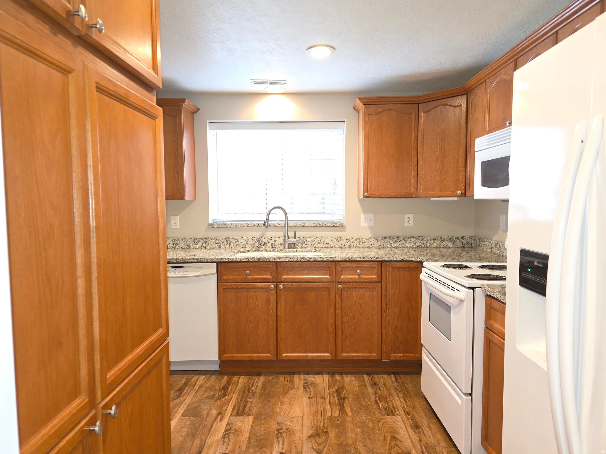 Efficient kitchen featuring wood-finish cabinetry, speckled countertops, stainless steel sink with gooseneck faucet, white appliances, and wood-finish flooring