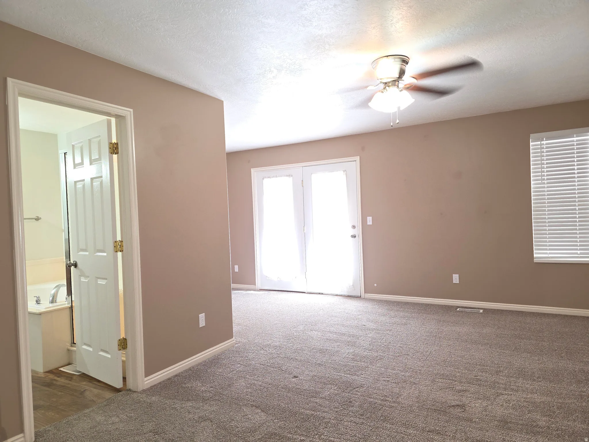 Spacious room featuring neutral carpeting, a ceiling fan, and double French doors with white sheer curtains