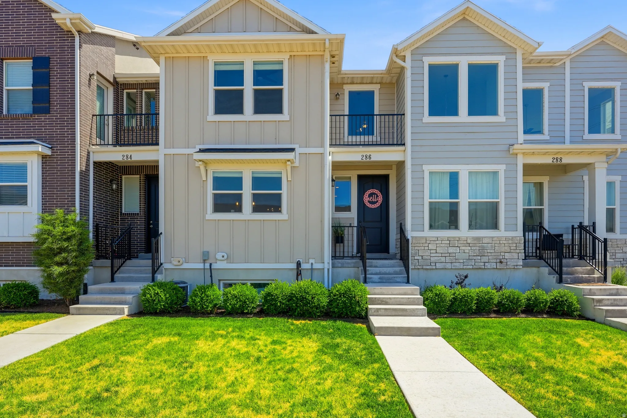 Contemporary townhome exterior featuring light beige vertical and horizontal siding, a stone accent facade, black window frames, and an upper-level balcony with black iron railings