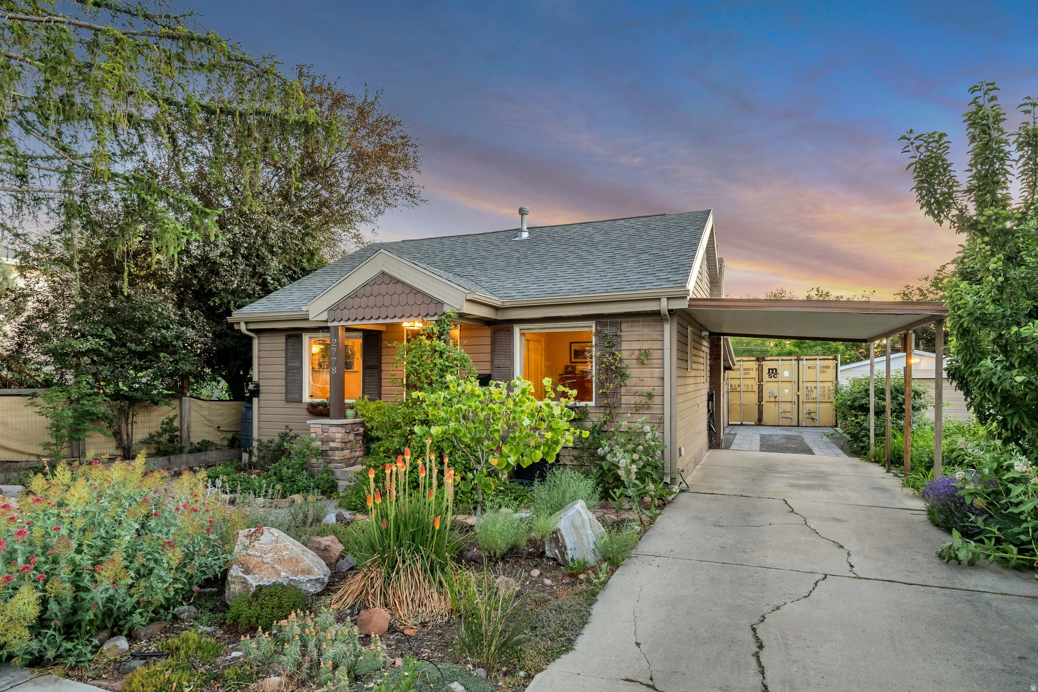Front elevation featuring brown siding, a covered entry with stone accents, a carport, and a mature landscaped garden with various plantings and rocks