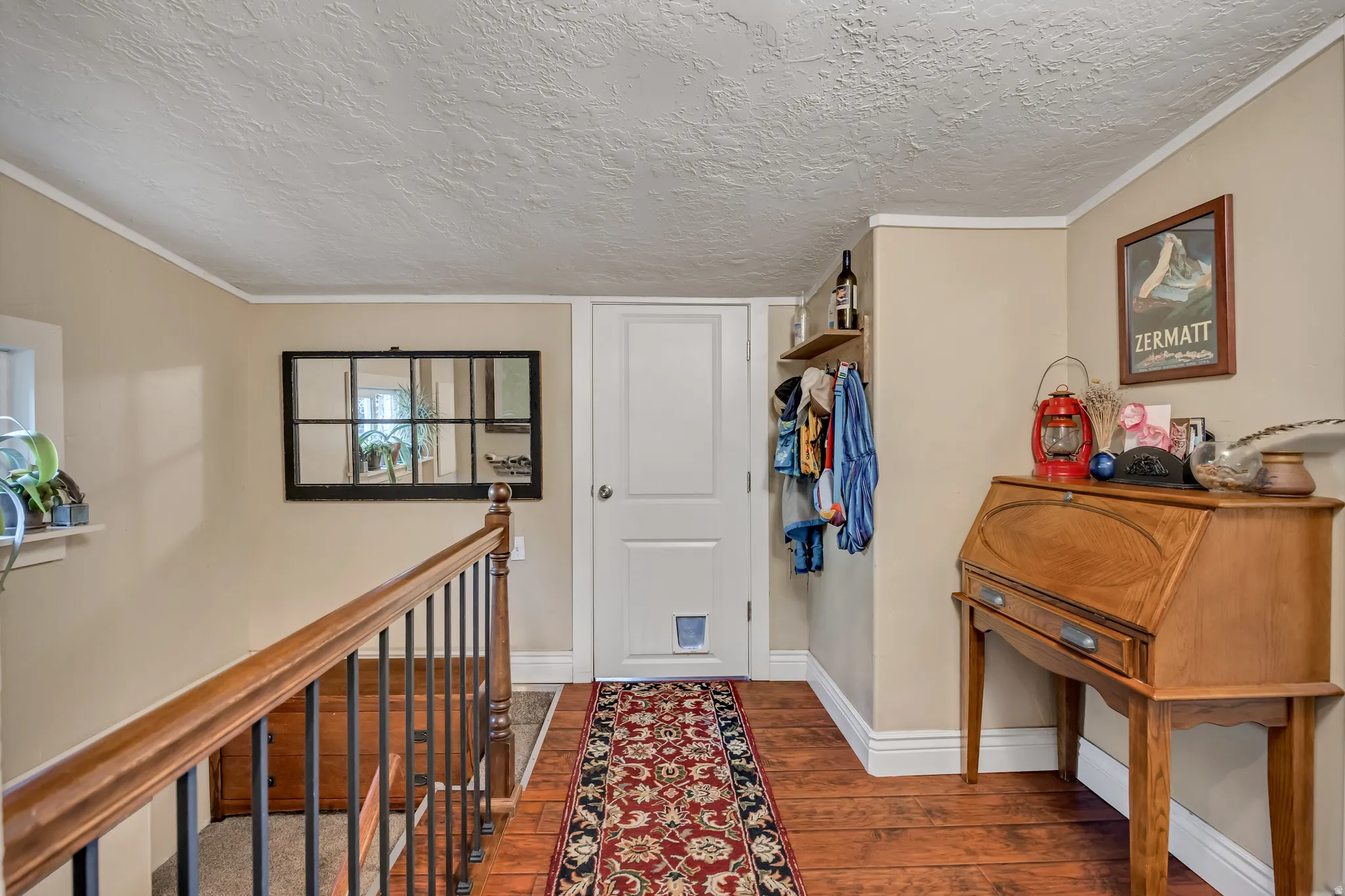 Wood-finish flooring with a textured ceiling finish, featuring a white paneled door, wood handrail with black iron balusters, and cream-colored walls with white trim