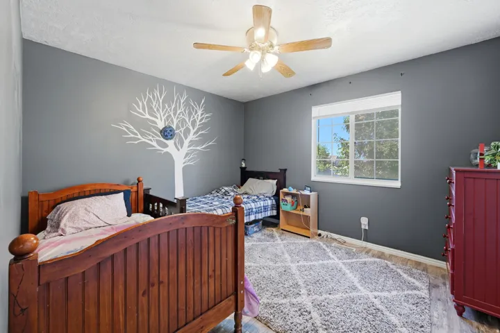 Spacious room featuring a ceiling fan with integrated lighting, a large window providing natural illumination, wood-finish flooring, and a decorative wall decal
