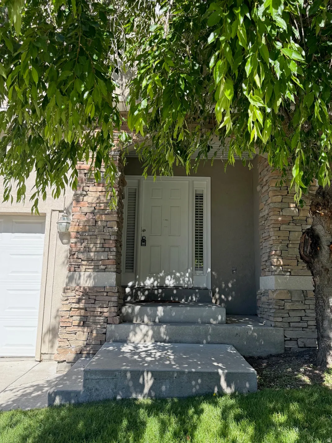 Front entry featuring a paneled door with sidelights, stacked stone accents on architectural columns, a stepped concrete walkway, and a garage with a white sectional door