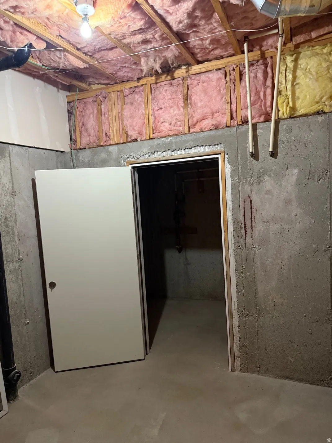 Unfinished basement area featuring exposed ceiling joists with insulation, concrete walls, and a solid core door