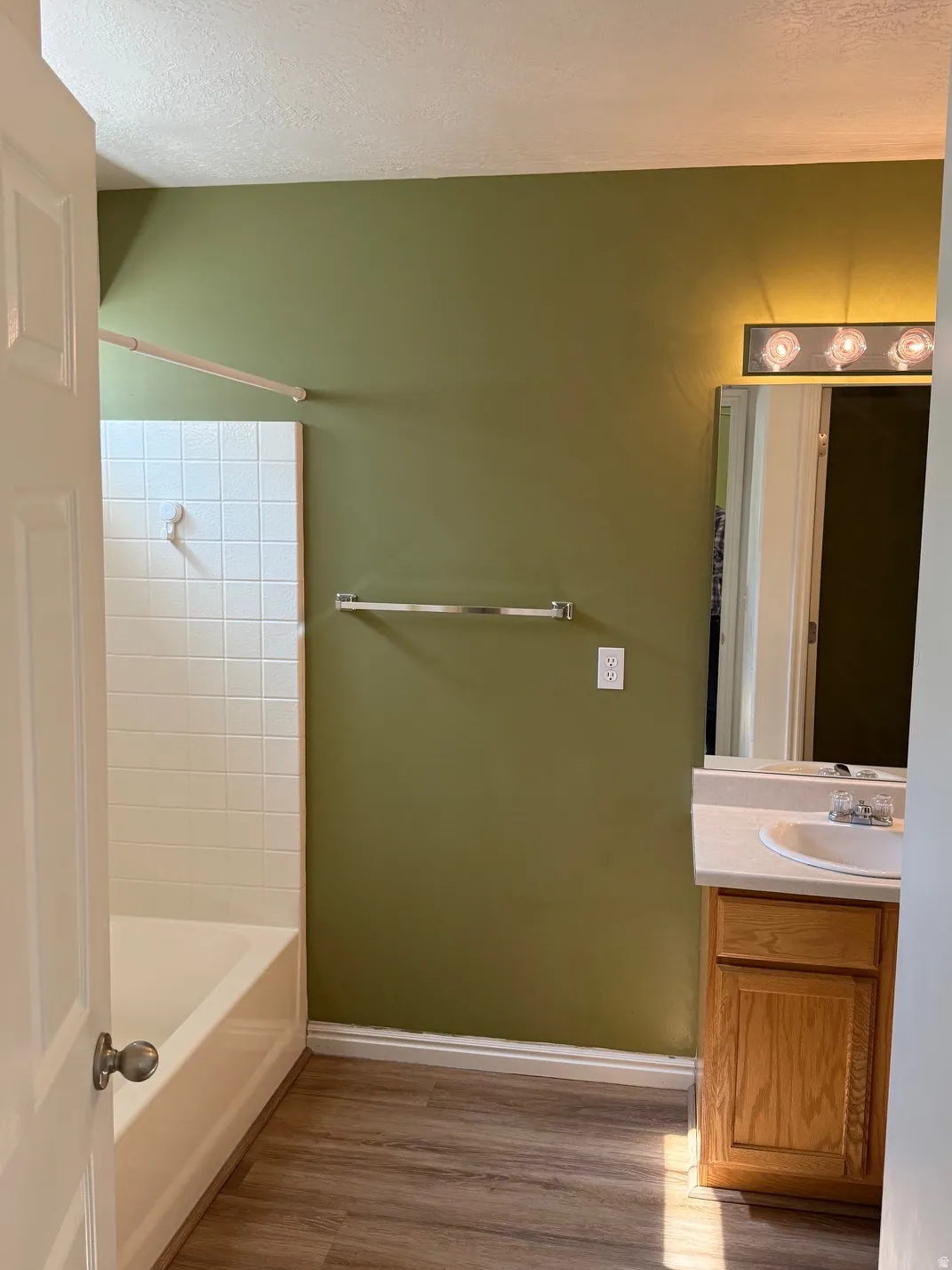 Master Bathroom featuring a white tub with subway tile surround, wood-finish flooring, single vanity with light-toned countertop, and a wall-mounted mirror with an overhead light fixture