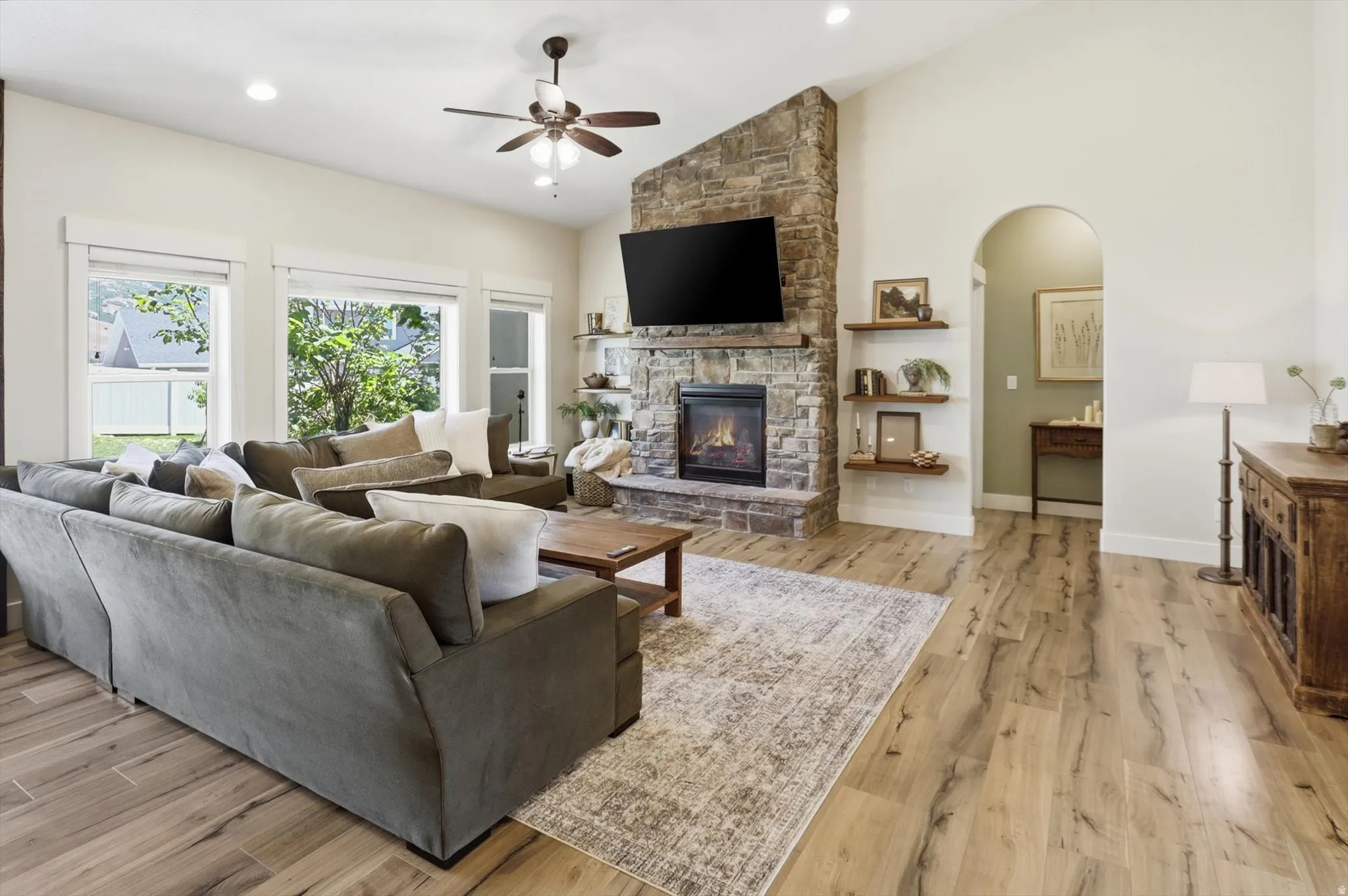 Spacious living area featuring wood-finish flooring, a vaulted ceiling, a stone-clad fireplace with an integrated mantel, and multiple windows