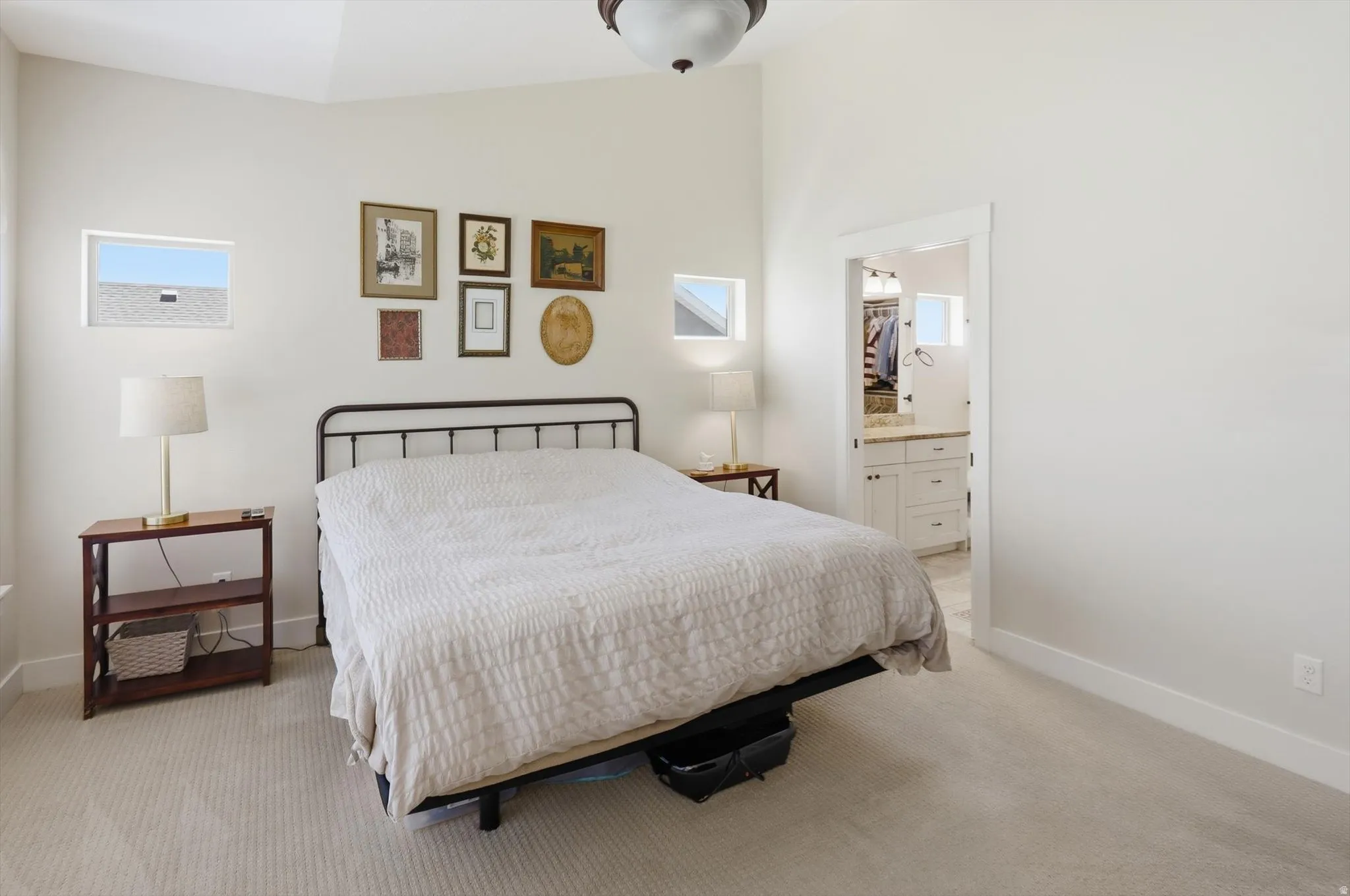 Spacious bedroom featuring vaulted ceilings, light-toned carpeting, and a doorway revealing a bathroom with a white vanity and stone countertop