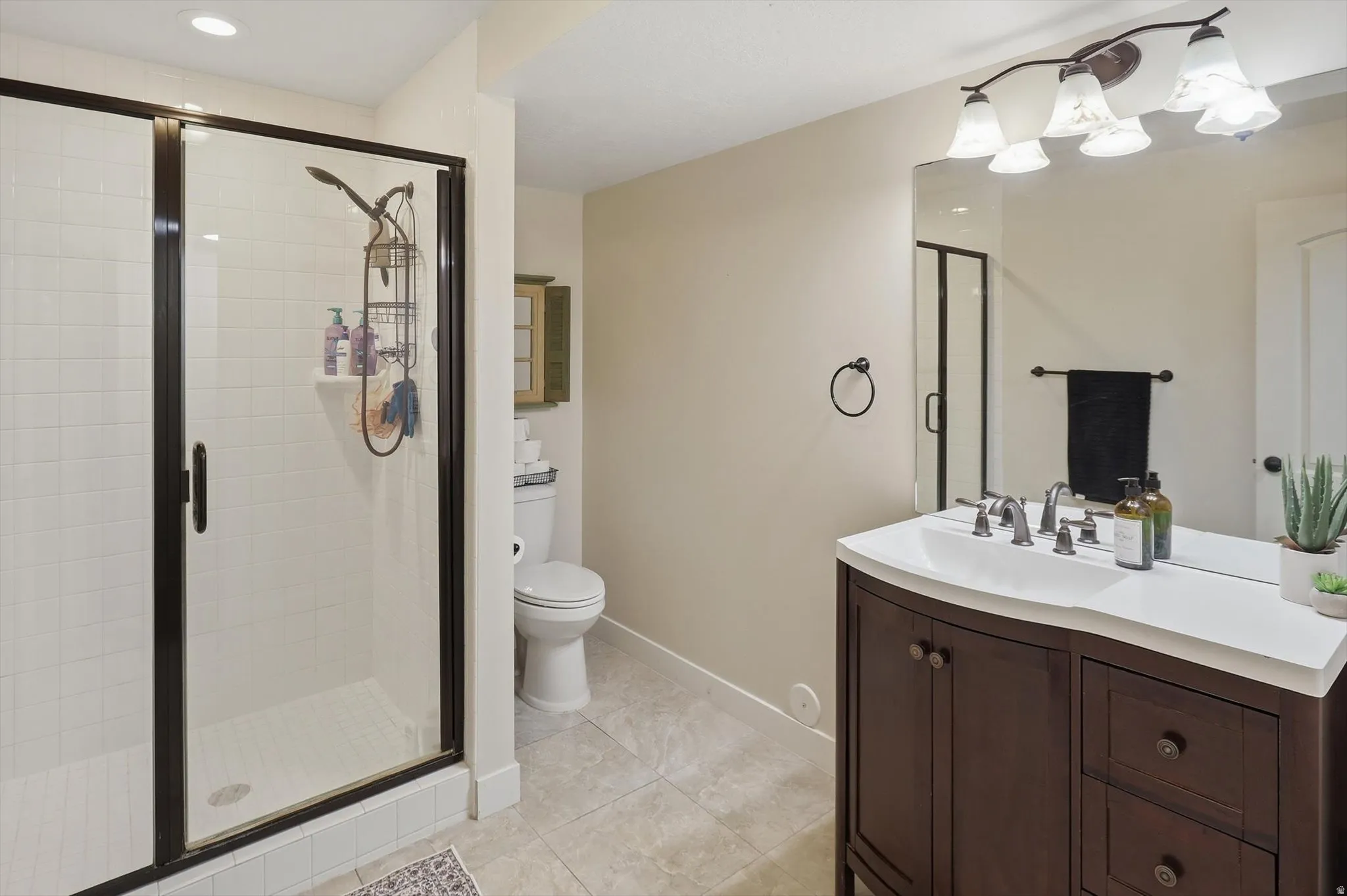 Bathroom featuring a dark wood-finish vanity with an integrated white sink and counter, a frameless mirror, a five-light fixture with frosted glass shades, a glass-enclosed shower with subway tile, and light-toned tile flooring