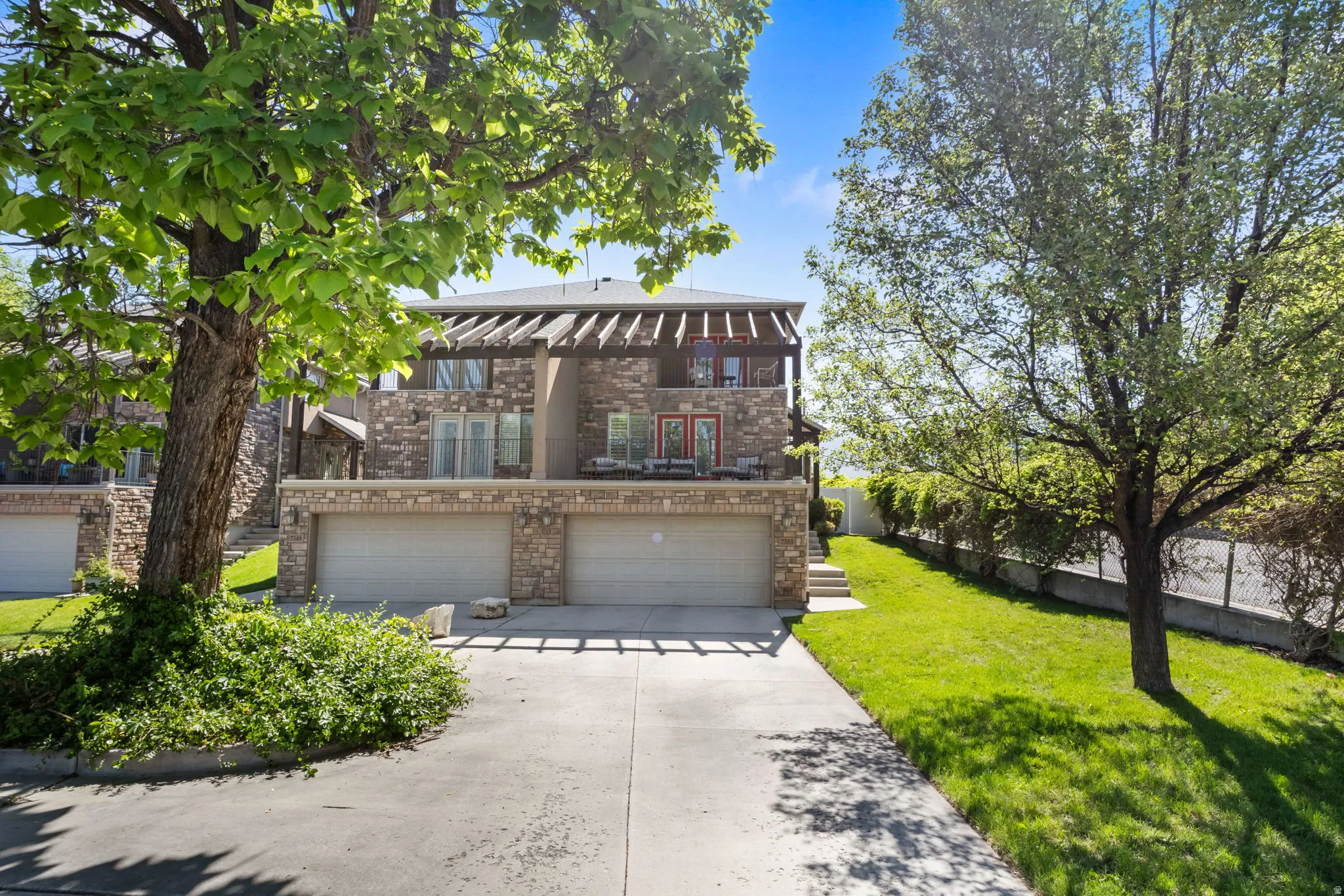 Stone-clad exterior featuring dual garages with overhead doors, a second-story balcony with pergola, and a vibrant green lawn